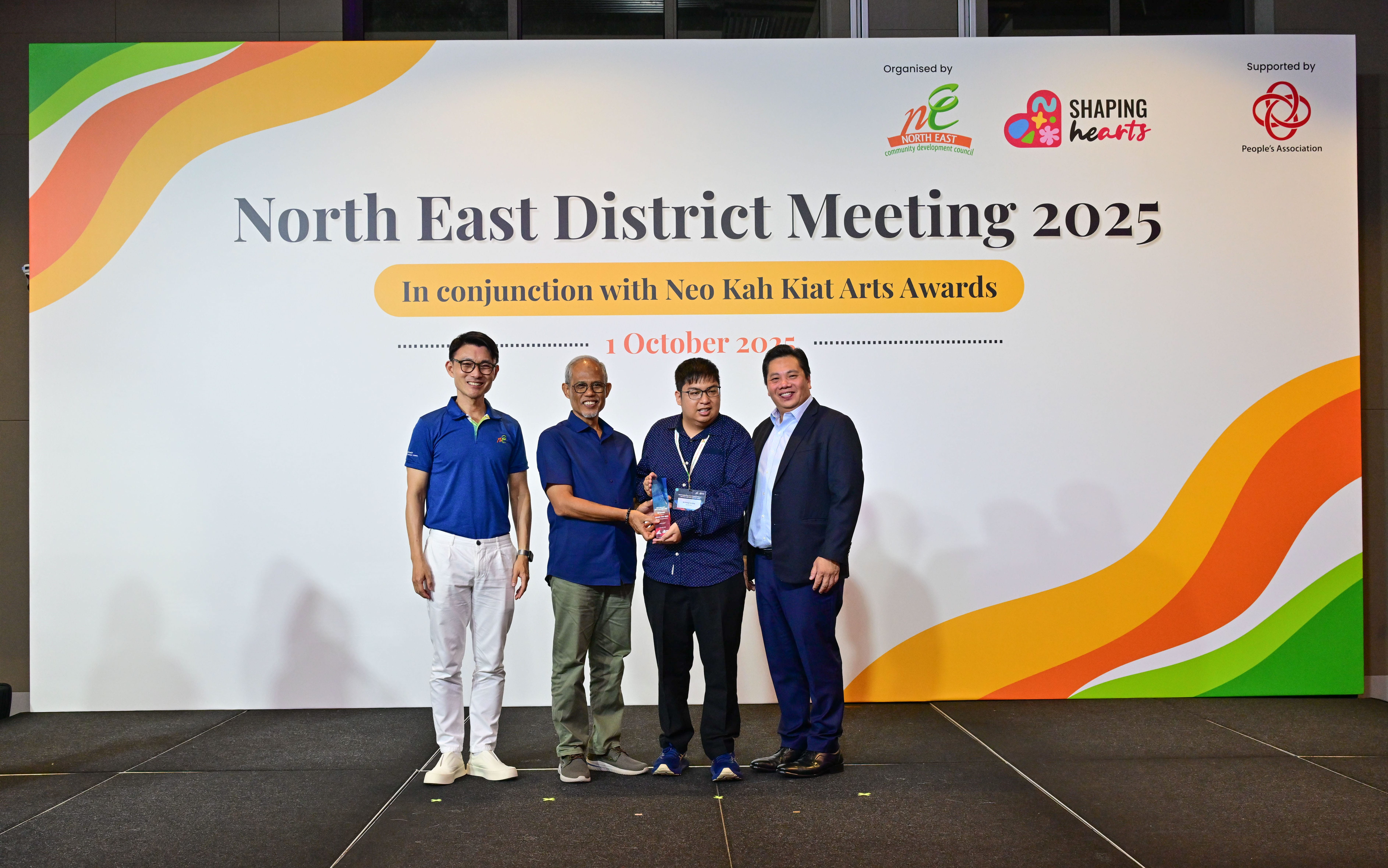Award recipient standing on stage with the presenter, holding a acrylic plaque and posing for a group photo during the award ceremony, with a backdrop reading ‘North East District Meeting 2025 In conjunction with Neo Kah Kiat Arts Awards'